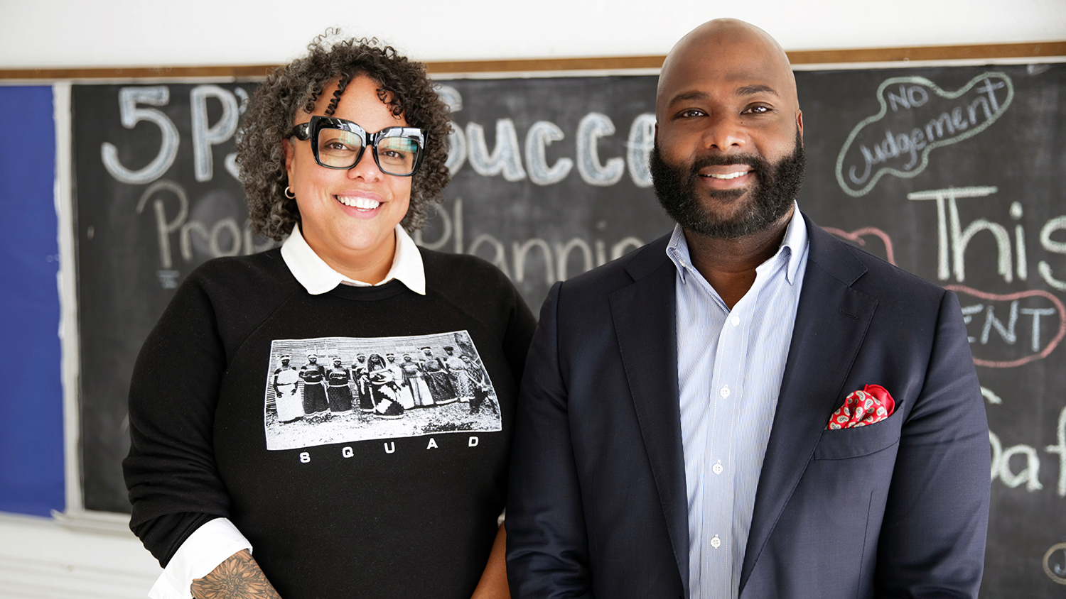 RICH co-founder Danielle Battle, wearing her trademark stylish eyeglasses and a dark sweatshirt with a photo of people and the word “Squad” on it, poses for a photo with Frederick Riley, executive director, Weave: The Social Fabric Project, who is in an open-collared shirt and a navy blazer. They are standing in front of a blackboard filled with inspirational phrases at RICH’s headquarters in Baltimore.  