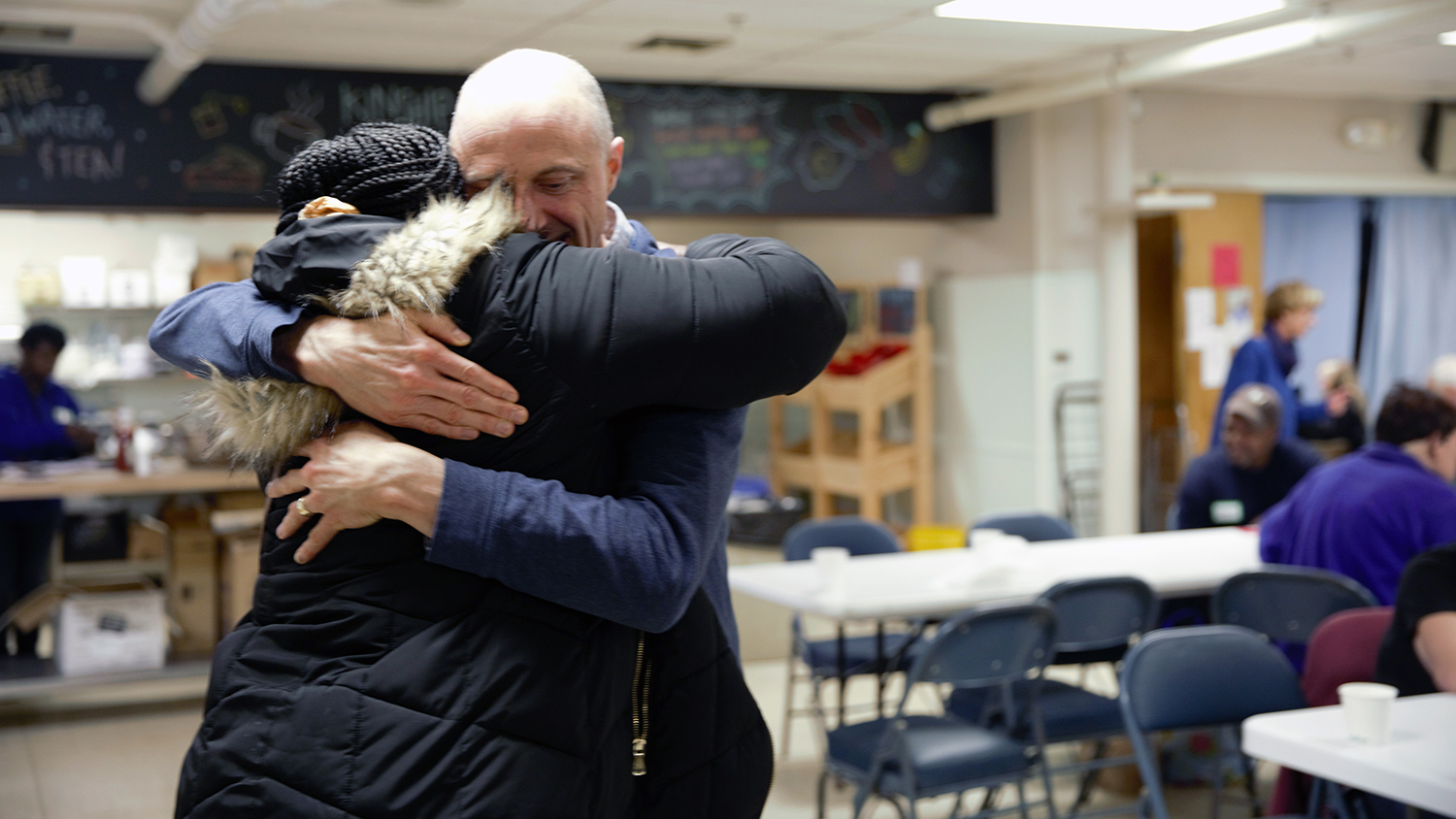 Vincent Noth, dressed in a dark sweater, hugs a woman in a heavy winter coat who is a community member at Kinship Community Food Center. The center is in the basement of St. Casmir Catholic Church, where dozens of people have gathered for dinner in a brightly lit room lined with tables and chairs.  