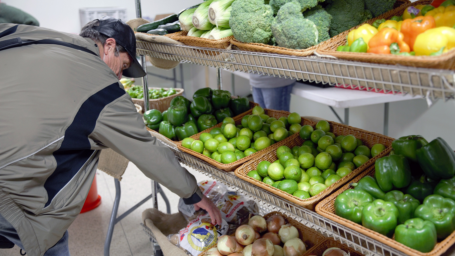 In the center’s market, a man in a gray jacket, who is a Kinship community member, selects onions from wicker baskets on long wire shelves filled with produce, including green peppers, limes, and broccoli.  