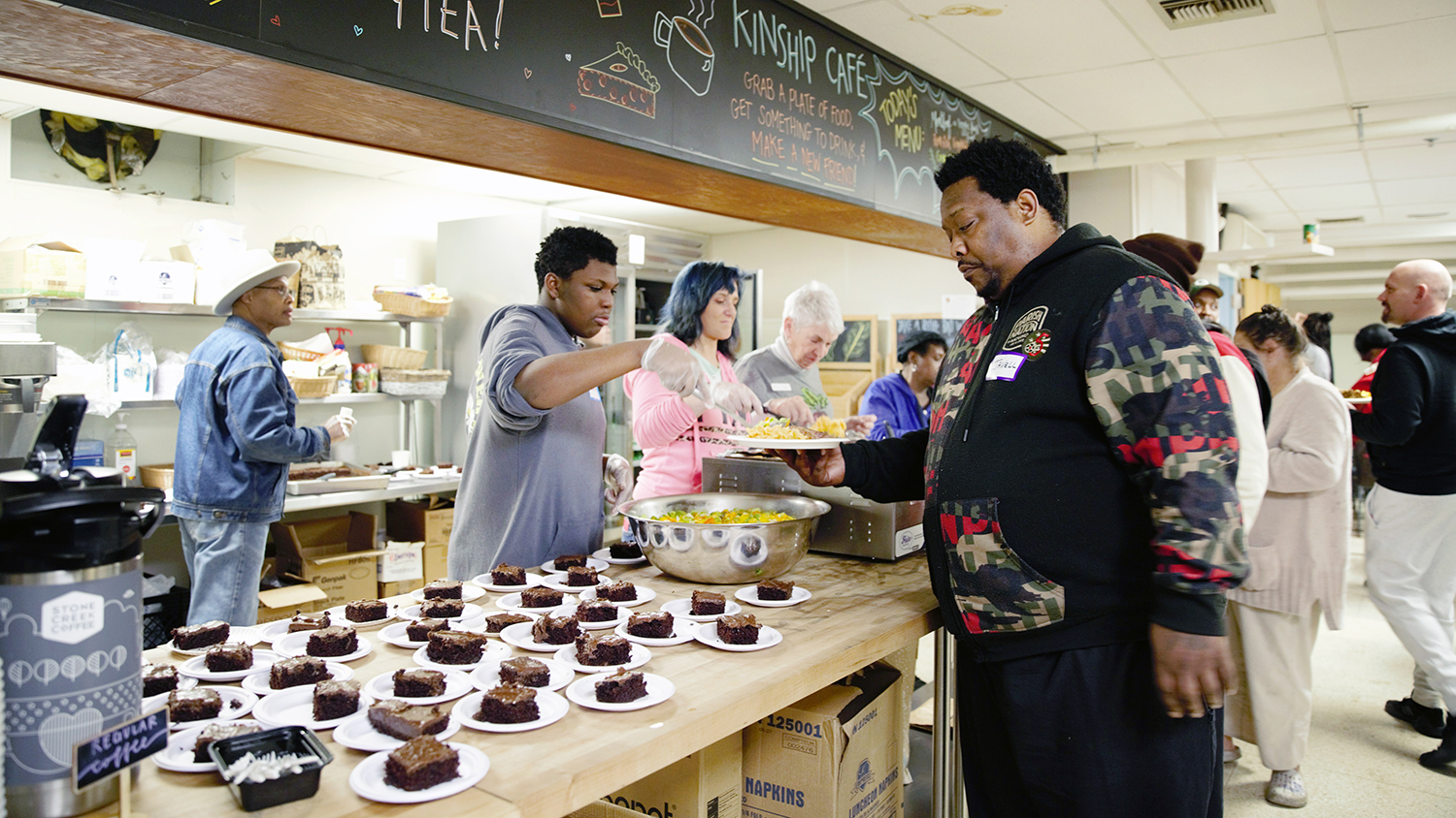 Under a blackboard with a colorful sign reading “Kinship Café,” Kinship volunteers spoon out food for community members who have lined up at a long counter that is also filled at one end with plates of pre-sliced peanut butter chocolate cake for dessert.  