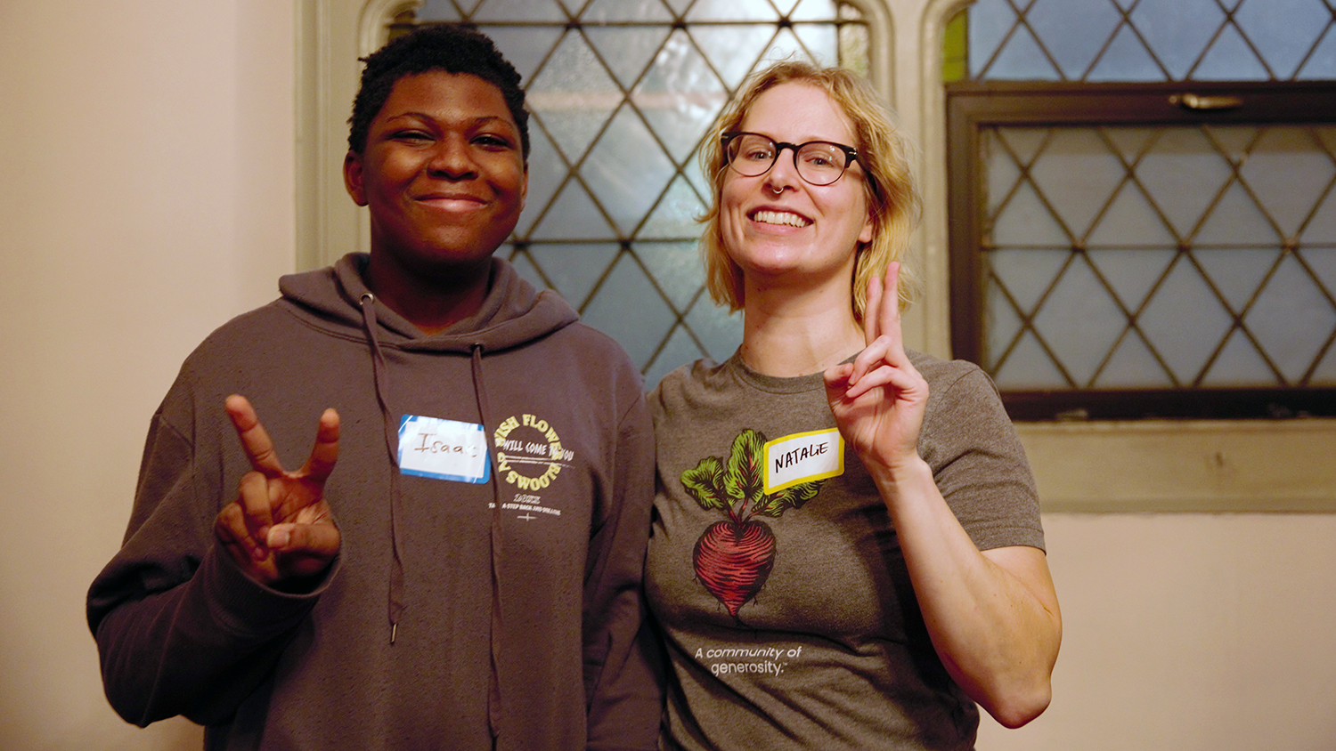 Isaac Williams and Natalie Ross stand side by side, each offering a smile and a peace sign to the camera. He is wearing a hooded sweatshirt, and she is wearing a Kinship T-shirt adorned with a radish and bearing the phrase “A community of generosity.” Like everyone at Kinship, they are each wearing name tags.  