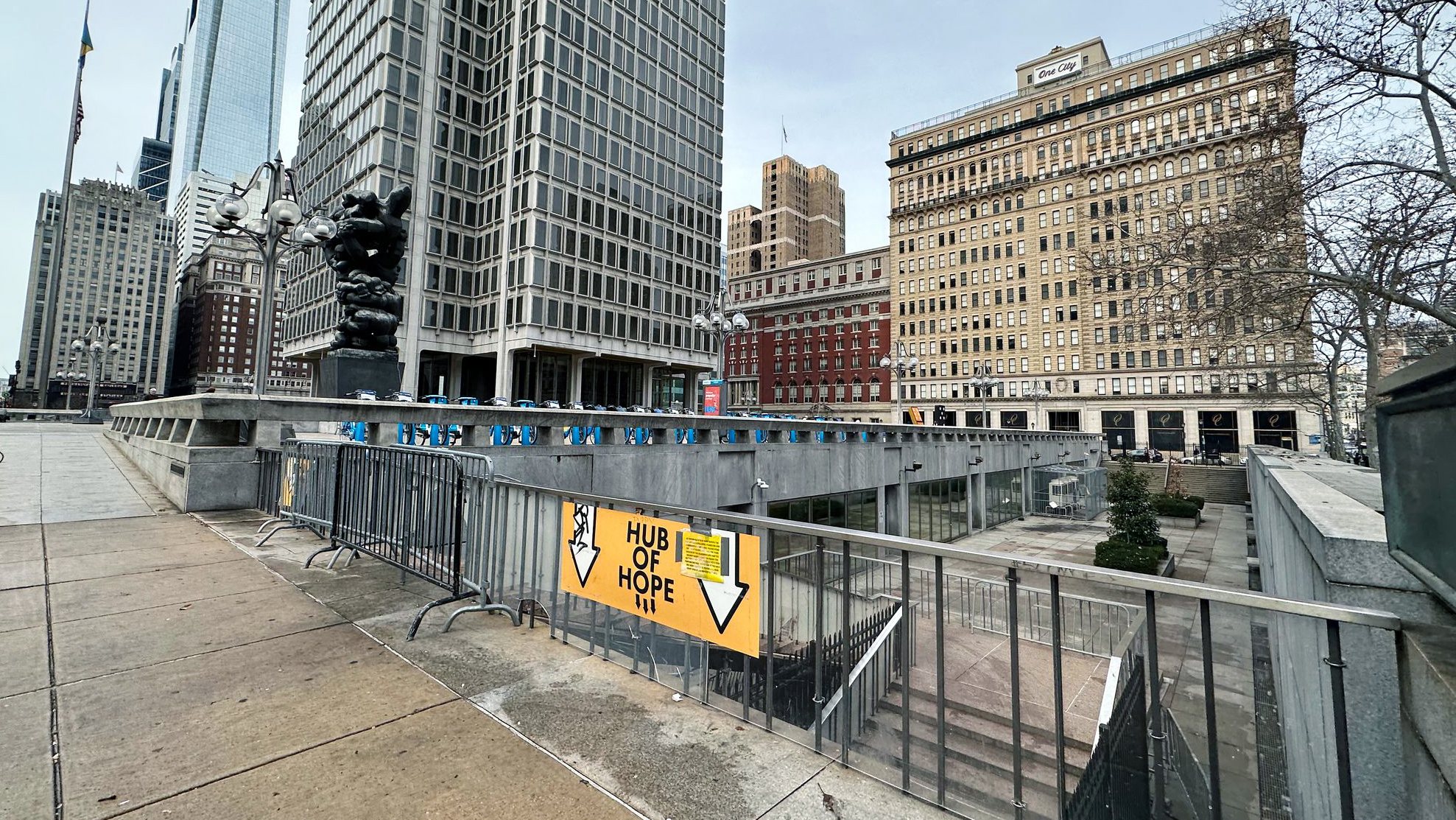 A yellow sign with multiple downward arrows indicates that the “Hub of Hope” is down steps to a former subway station below the downtown streets. A large glass skyscraper and other tall buildings are in the background against a clear but gray sky. 