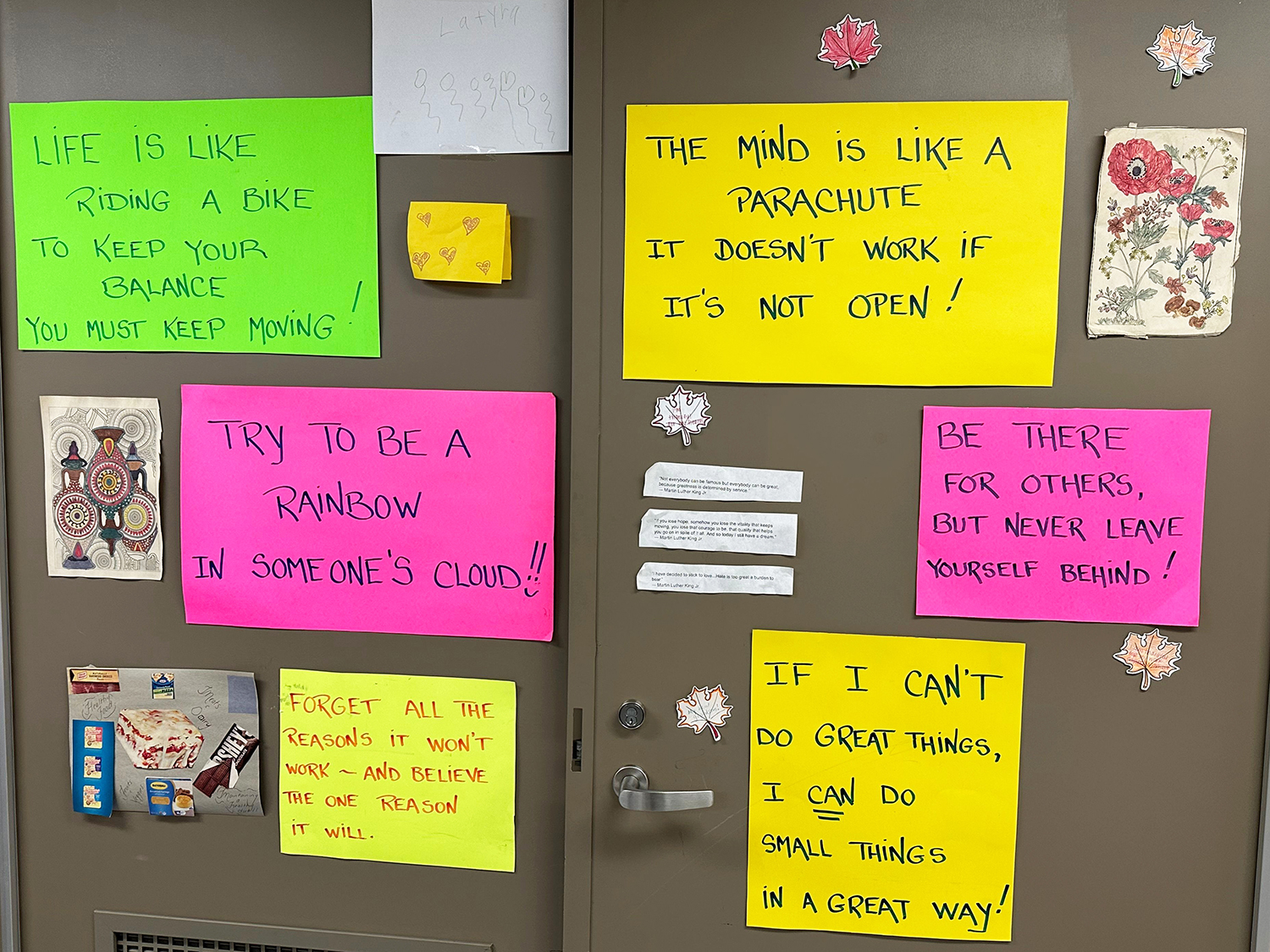 Several inspirational phrases are written on yellow, pink, and green sheets of paper, adding a colorful touch to a set of grayish-brown double doors. The sayings include, “Try to be a rainbow in someone’s cloud,” “Forget all the reasons it won’t work—and believe the one reason it will,” and “Be there for others, but never leave yourself behind.”