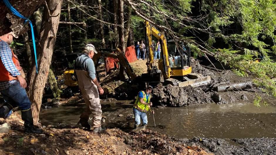 Three workers, wearing waders or high boots, stand on the muddy banks of the brook. A yellow piece of construction machinery sits on the opposite bank.  