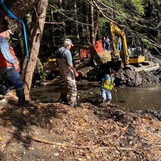 Three workers, wearing waders or high boots, stand on the muddy banks of the brook. A yellow piece of construction machinery sits on the opposite bank.  