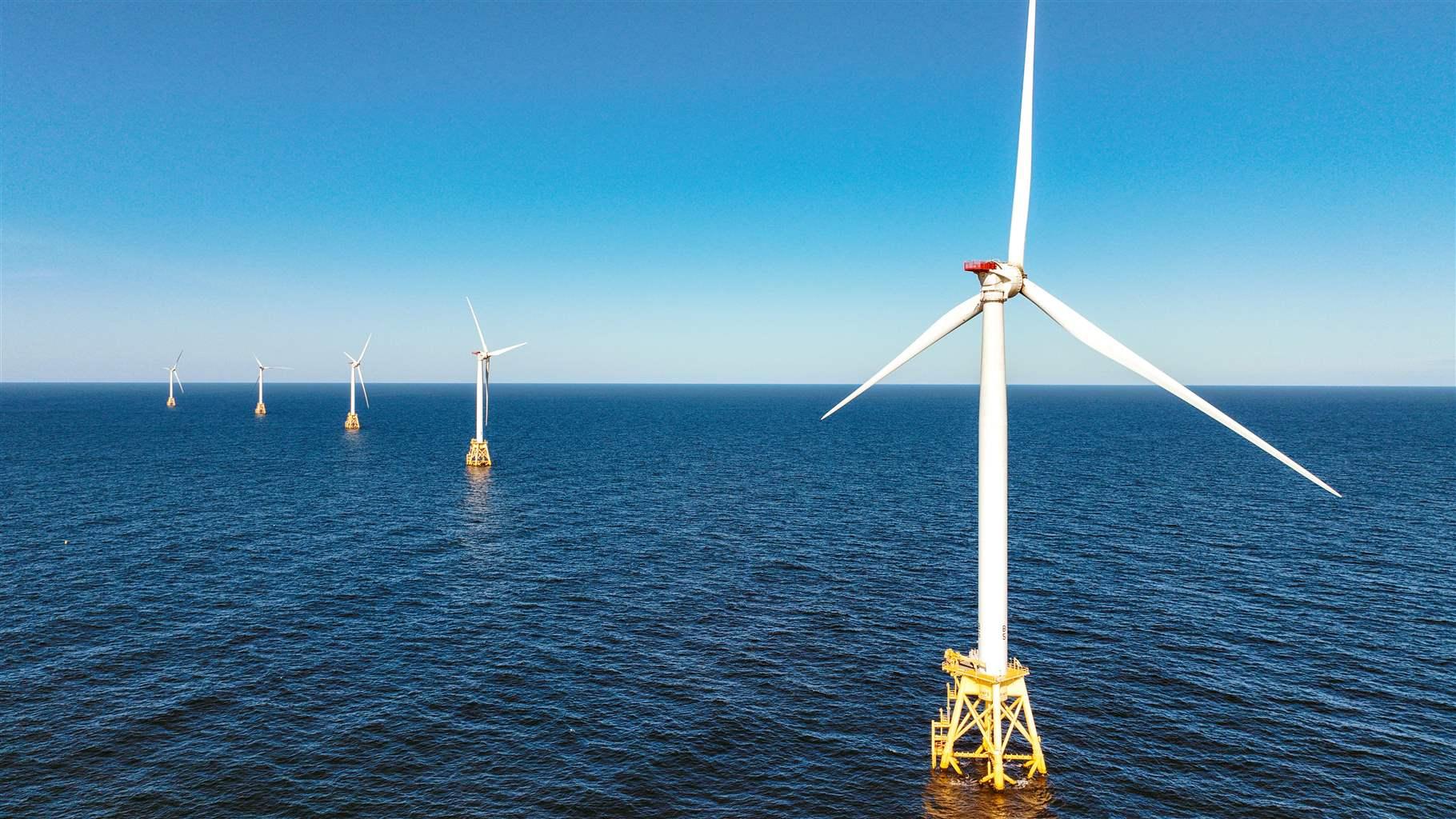  Five windmills mounted on small, yellow platforms run diagonally out to a light blue horizon where the sky meets the darker blue ocean. 