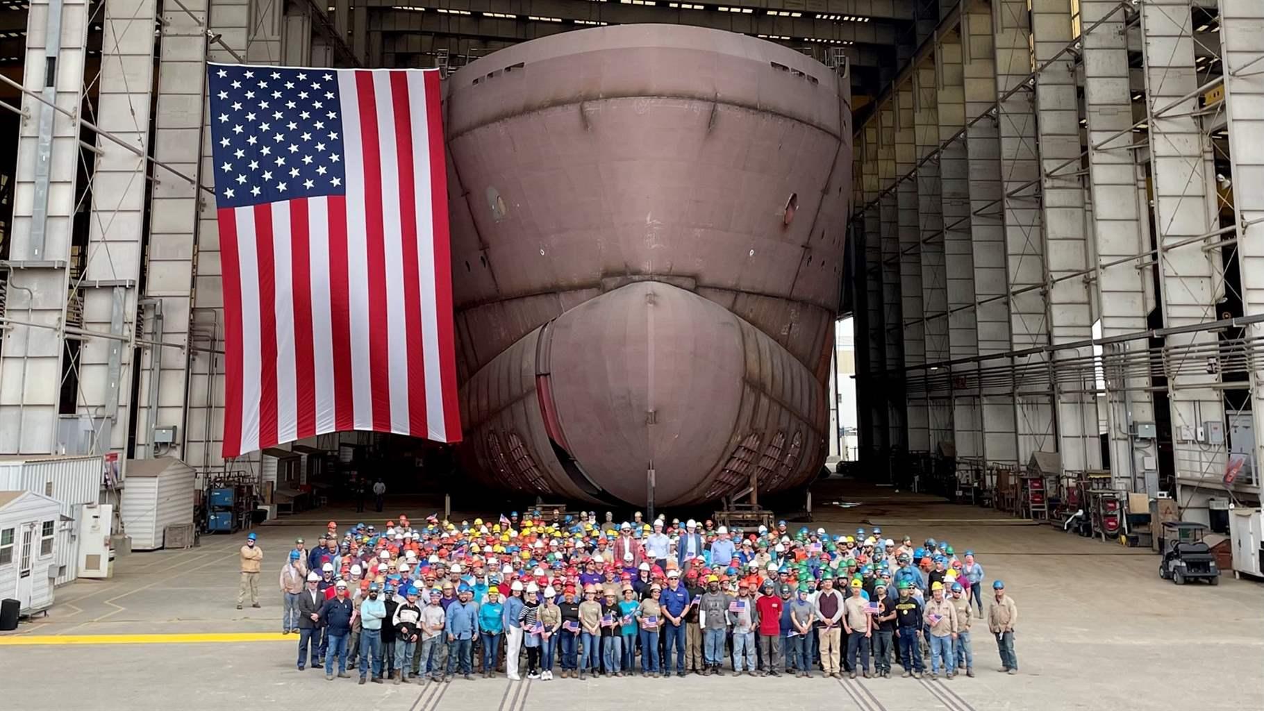 A group of at least 100 people stand on a concrete dry dock in front of the bow of a huge ship that they are helping to build. Massive scaffolding rises on both sides of the boat and extends over its top, and a large American flag hangs from that scaffolding next to the ship’s bow.