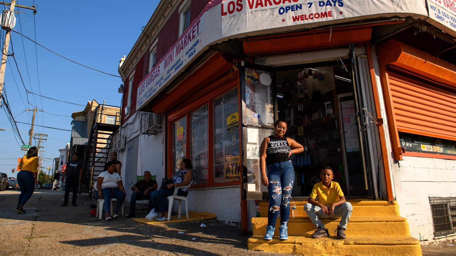 A girl and a boy pose on the bright yellow steps of a corner store under a sign that reads: “Los Vargas Mini Market, Open 7 Days, Welcome.” To their left, in the background, a small group of adults gather, some seated in patio chairs and some standing. 