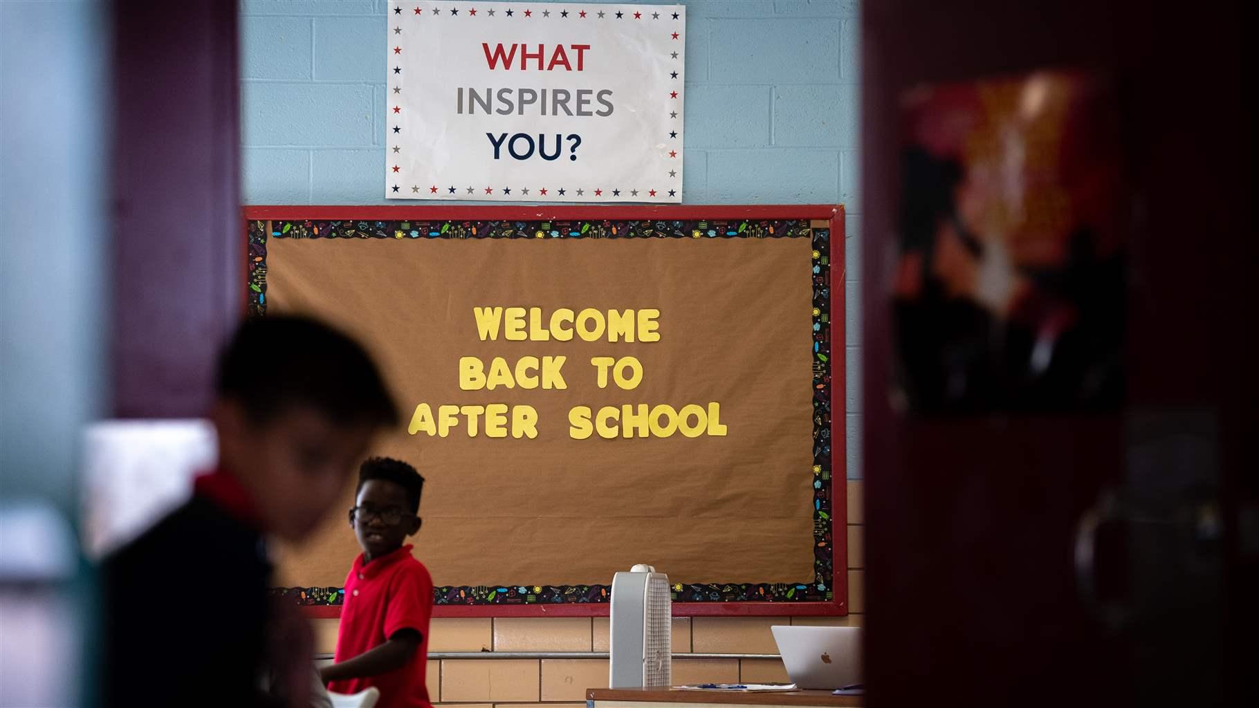 Yellow cutout letters on a bulletin board spell: “Welcome Back to After School.” Above that, a white sign with red, blue, and gray letters reads, “What Inspires You?” An elementary school-age child stands in front of the signs, while another stands out of focus closer to the camera.