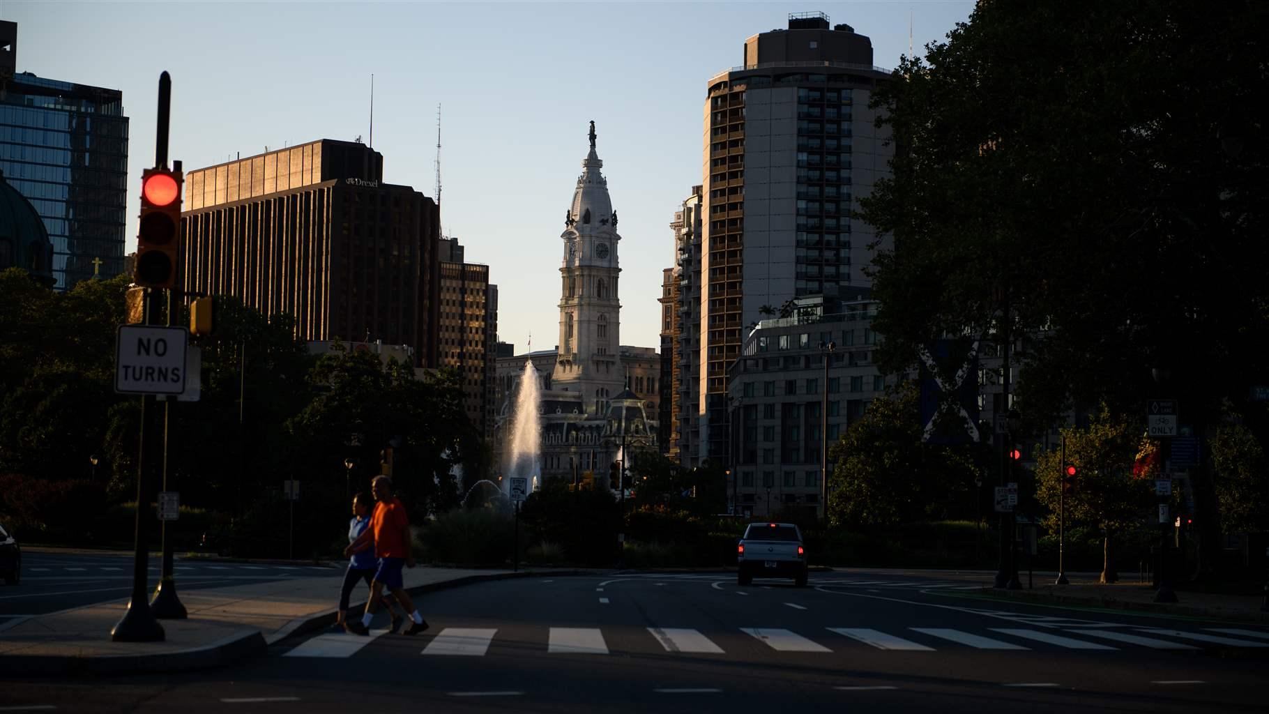 A plume of water from Swann Memorial Fountain sprays skyward in front of City Hall’s central tower, which sits in the middle of several tall downtown buildings. Two pedestrians in casual attire walk on a crosswalk in heavy shadow in the foreground.