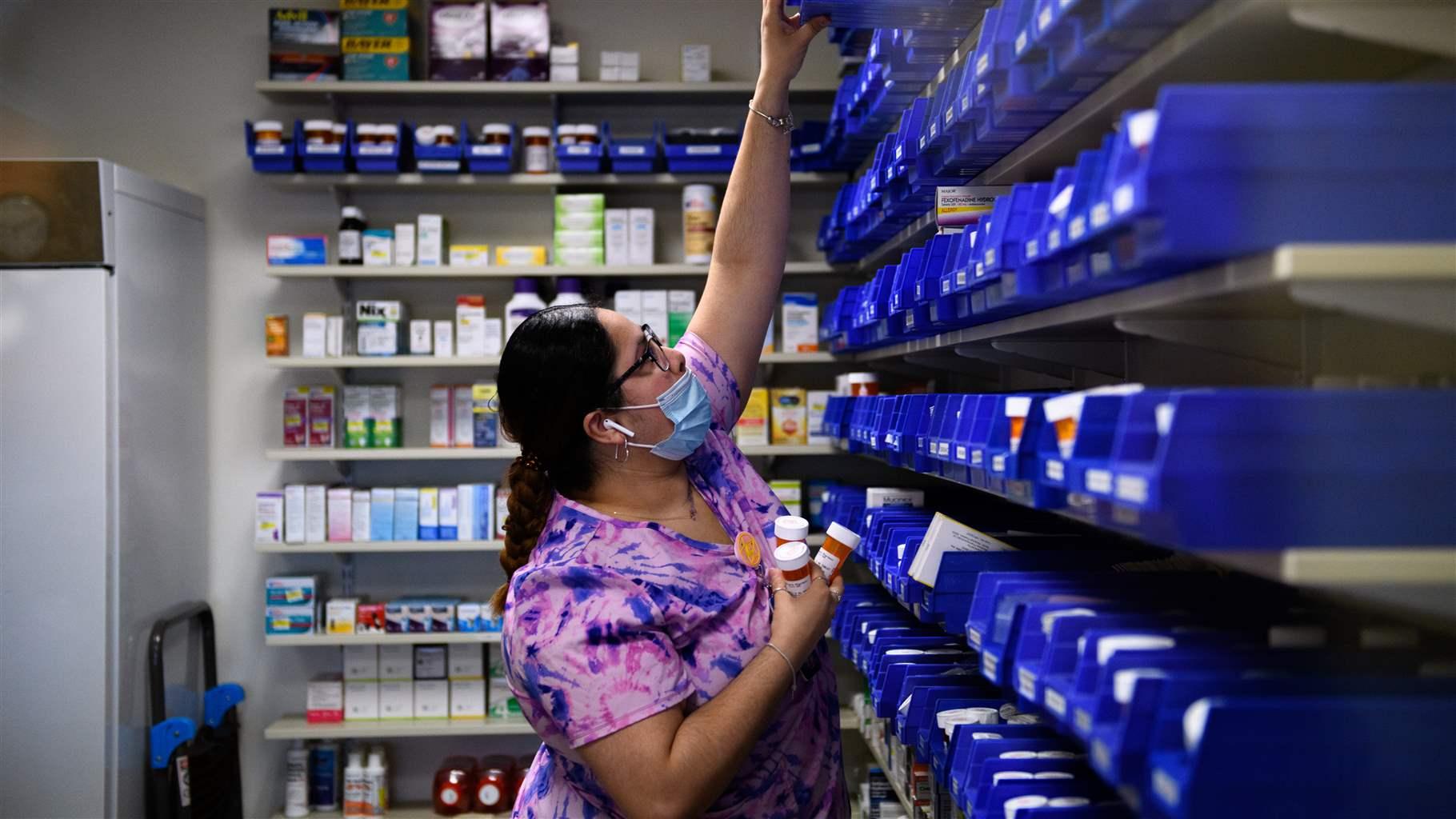 A woman wearing a light-blue surgical mask, glasses, and a purple-and-blue shirt reaches high for a blue plastic container in a room lined with medication-filled pharmacy shelves. She holds prescription pill bottles in her other hand. 