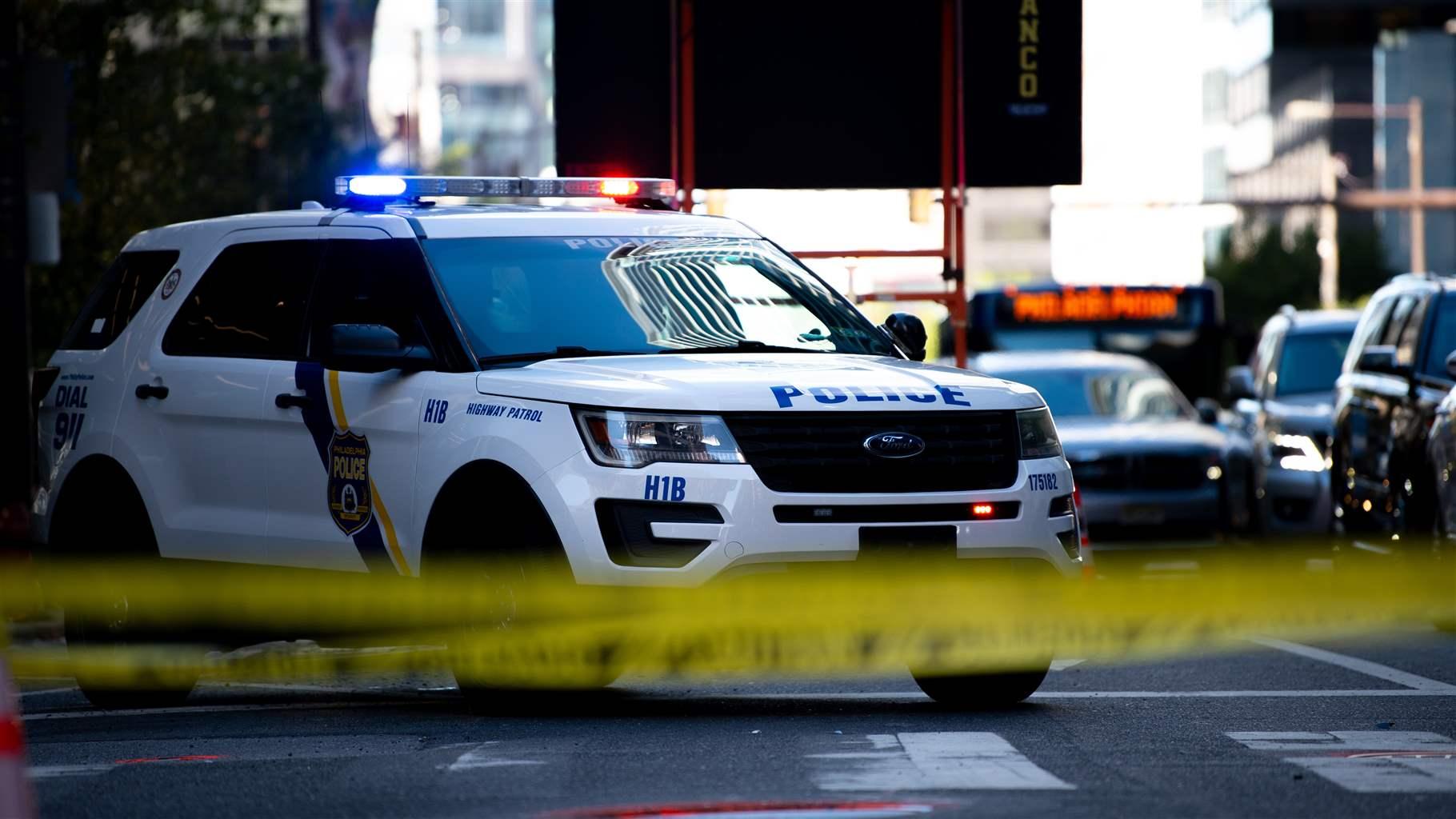 A white police SUV with its lights on is stopped in the middle of a city street. Blurred yellow police tape stretches across the front of the photo. 