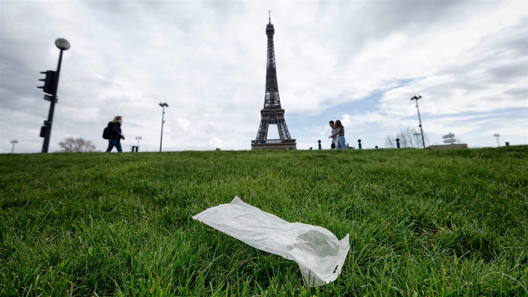A white, medium-sized plastic bag lies on a thick lawn of grass. In the background is the Eiffel Tower, a number of lampposts, and three pedestrians passing by.
