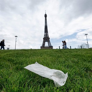 A white, medium-sized plastic bag lies on a thick lawn of grass. In the background is the Eiffel Tower, a number of lampposts, and three pedestrians passing by.