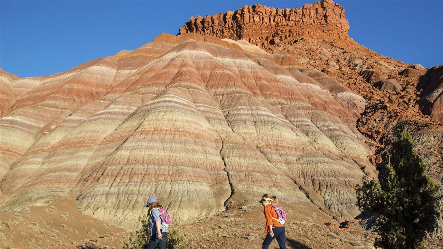 Two White women in comfortable hiking clothes walk on red earth past a large rock formation striated in the pastel browns, beiges, and reds of the U.S. desert Southwest. Beyond the formation, a burnt-orange mesa rises against a deep blue sky. Some native grasses and shrubs are present.