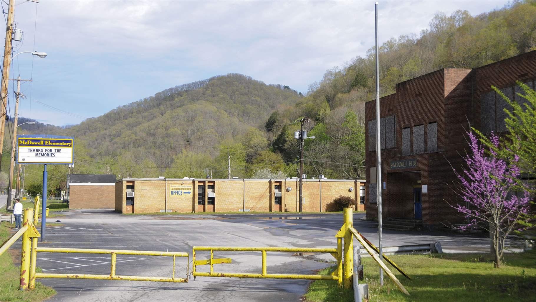 An asphalt driveway, blocked by a low-slung yellow gate on hinges, leads into an empty parking lot. On the right side is a two-story, red brick building with plywood over the windows. At the far end of the lot is a one-story brick building. A sign on a tall blue metal pole in the lot reads “McDowell Elementary: Thanks for the memories.” Forested hills rise behind both buildings.