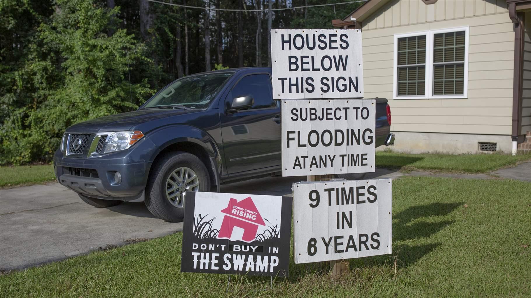 Four signs are staked into a lawn in front of a blue pickup truck and tan house. Three of the signs are stacked one on top of the other and read in bold black capital letters on white backgrounds: “Houses below this sign subject to flooding at any time. 9 times in 6 years.” To the left, the fourth sign shows an image of a tilted house sinking into a body of water surrounded by tall grasses and reads: “Horry County Rising. Don’t buy in the swamp.” 