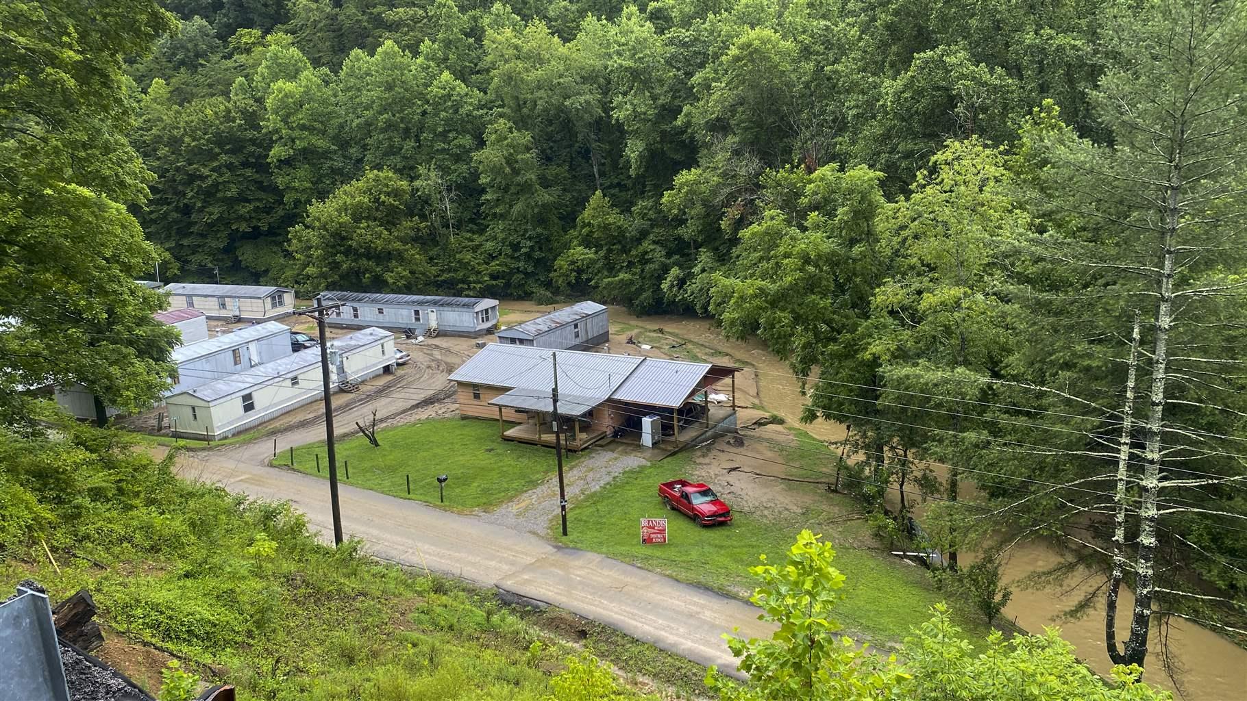 A view down into a mobile home park in a small valley. Half a dozen mobile homes sit close to each other, while a creek runs along one side of the park. The water in the creek is high and brown. A forested area sits opposite the creek from the mobile home park