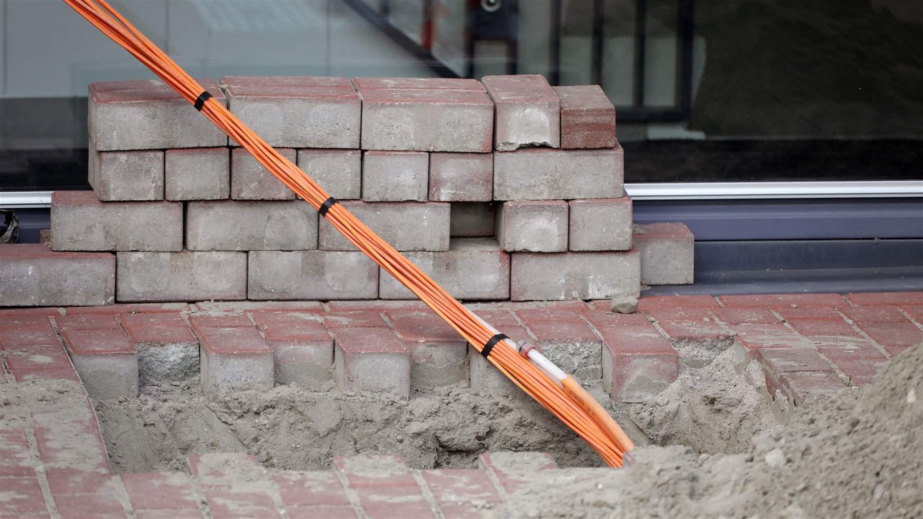 A long bundle of bright orange fiber optic cable sticks out of a hole in a partially constructed brick sidewalk. Mortar is also visible in the hole; a stack of bricks sits at the back of the sidewalk, and dirt is piled in the bottom right corner of the image.