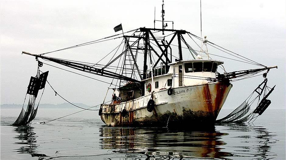 A white fishing vessel, heavily covered in rust and with extensive rigging, including nets draped into the sea, idles on a glassy ocean.