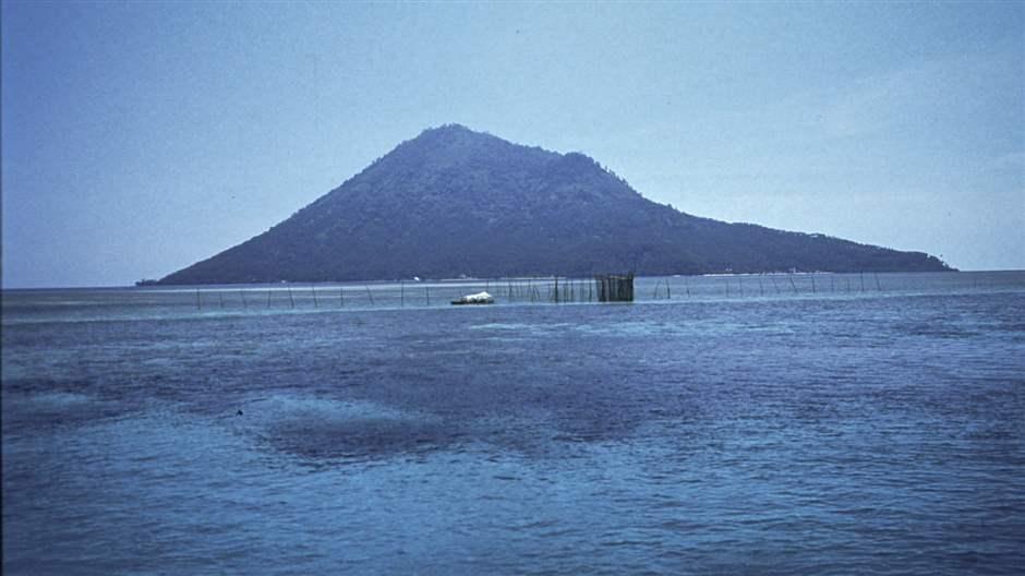 A mountain in the distance rises from a calm, tropical sea, and both land and water appear blue under a cloudless sky. In the middleground, a small fish trap made of sticks protrudes from the water.