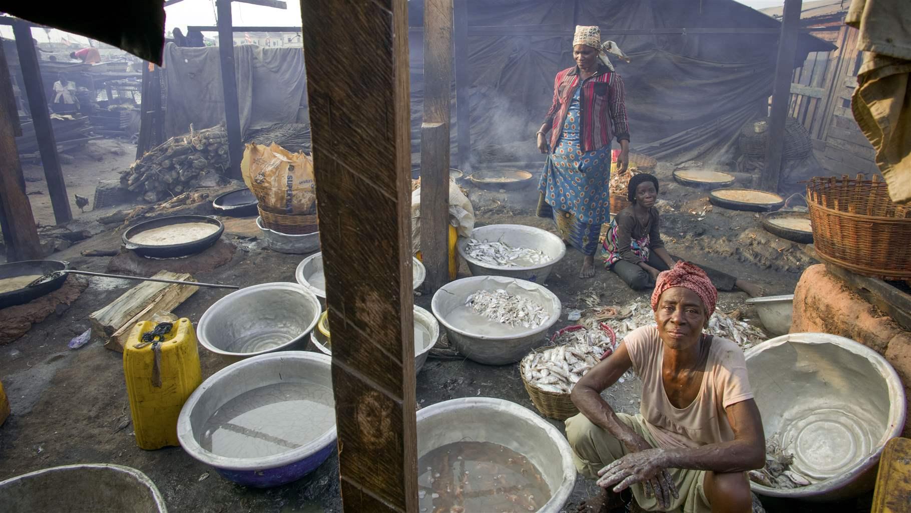 Three Black women—two seated and one standing—are in an open-air, dirt-floor area surrounded by metal tin basins, most empty but some with small dead fish in them. The air is smoky and the beams and frame of a roofless structure are visible around and above the women.