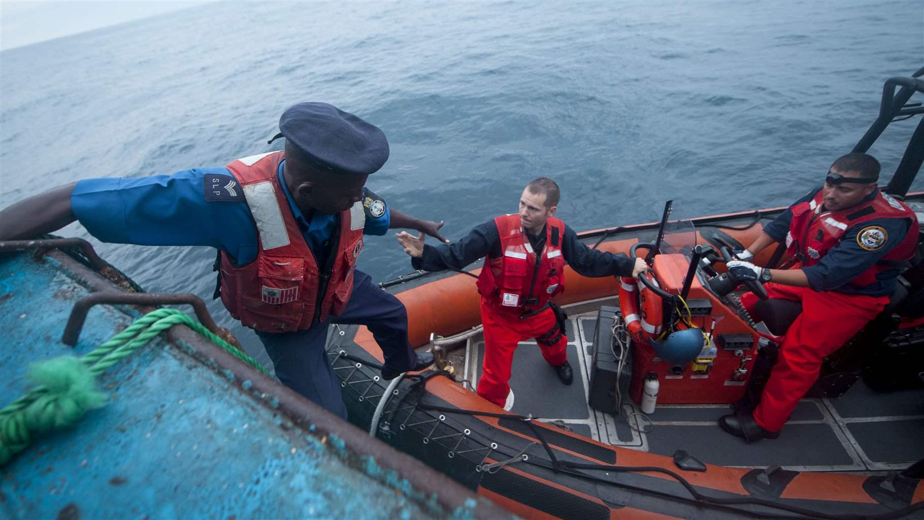 A Black man in military uniform descends from the blue deck of what appears to be a large boat into a smaller, orange, rubber boat, where a White man is extending a hand to help. Another Black man is at the wheel of the orange boat. All three men are wearing life jackets. 