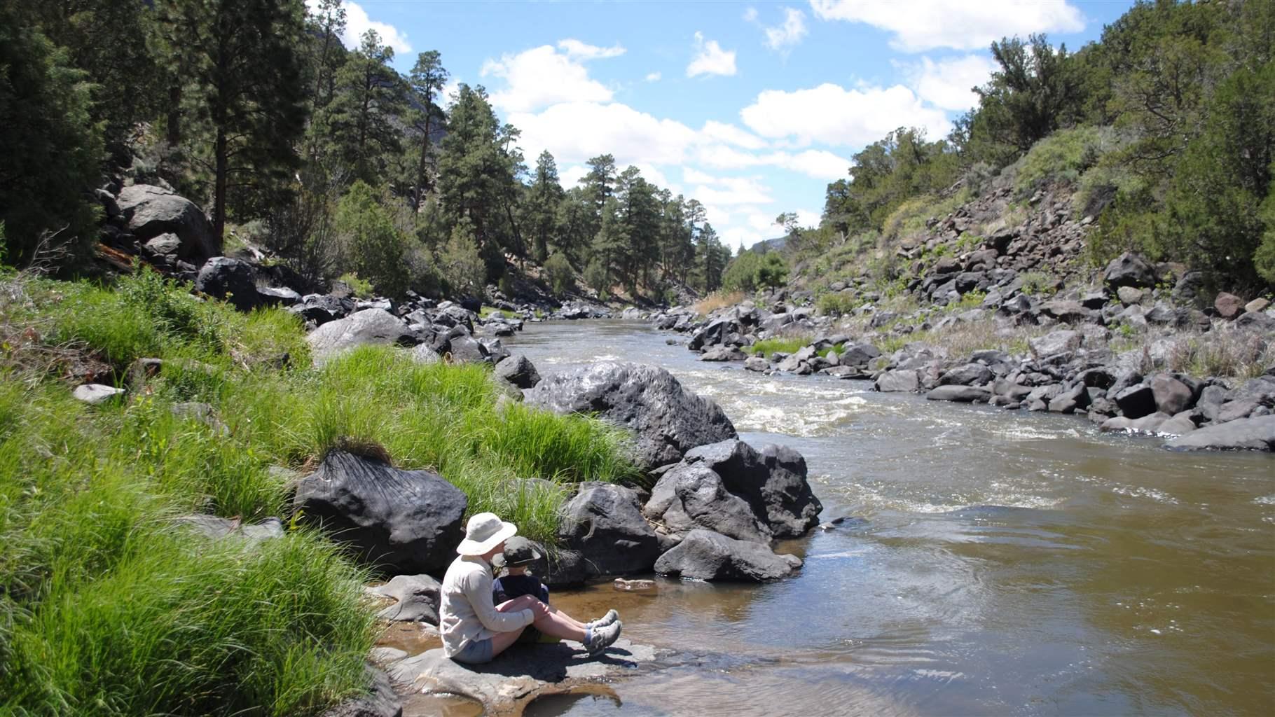 An adult and child, both in short pants, long-sleeve shirts, and sun hats, sit on a flat rock at the edge of a river. On both banks, rock-lined shores rise to bright green vegetation and, beyond, steep hills of conifer forests.