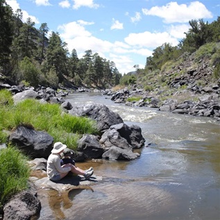 An adult and child, both in short pants, long-sleeve shirts, and sun hats, sit on a flat rock at the edge of a river. On both banks, rock-lined shores rise to bright green vegetation and, beyond, steep hills of conifer forests.