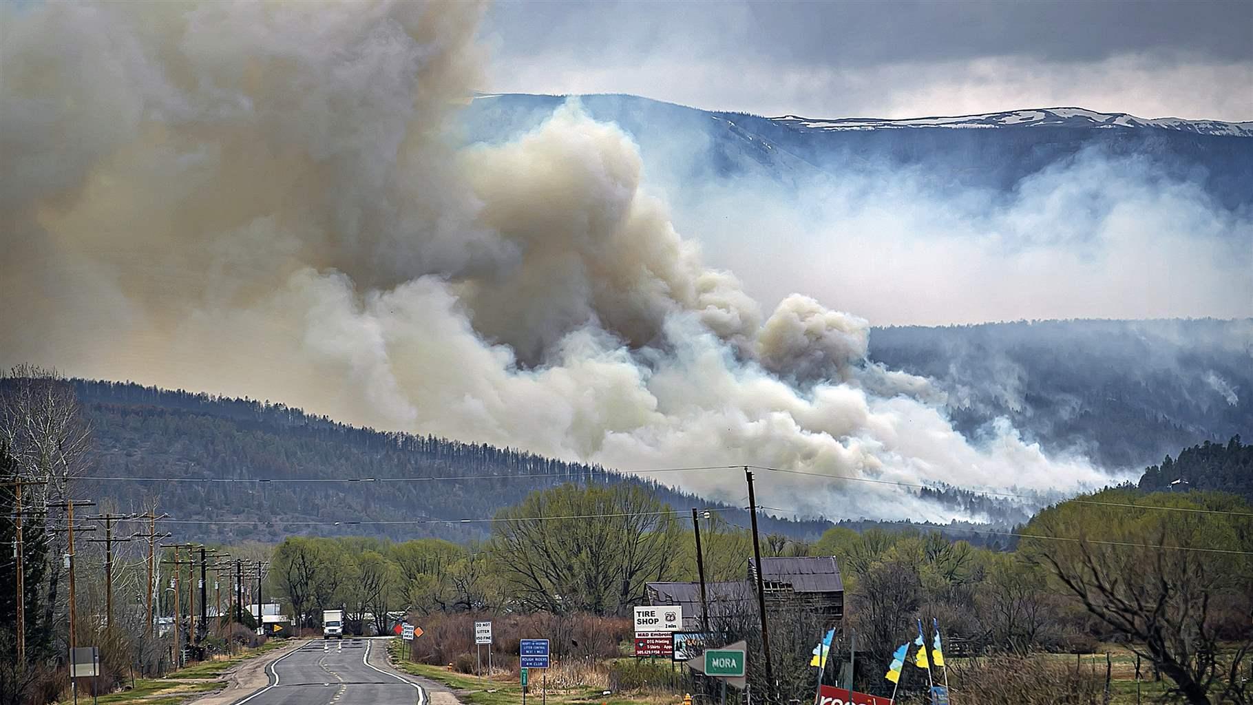 A two-lane highway stretches ahead toward forested foothills, behind which snow can be seen on the peaks of the crest of a mountain range. Clouds of smoke rise from the foothills, filling much of the sky. 