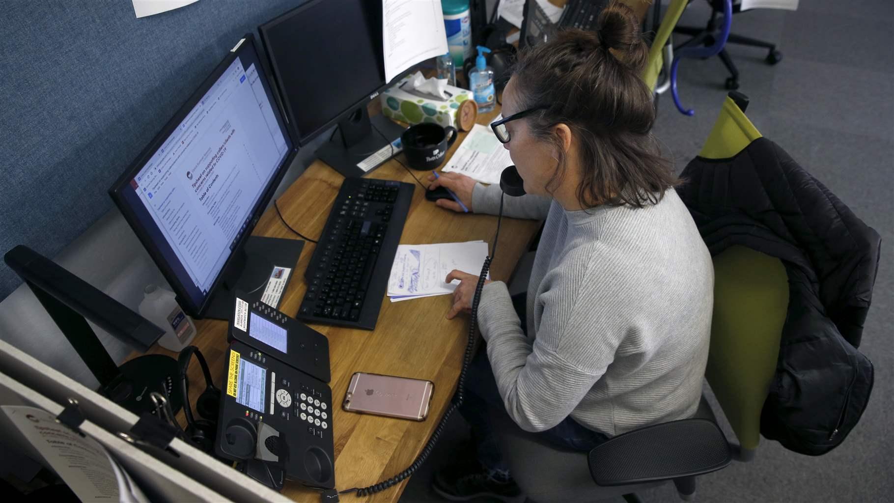A person sitting in front of a computer in an office talks on the telephone and tracks notes on papers on the desk. 