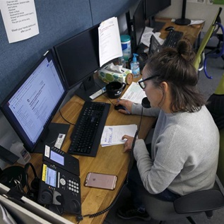 A person sitting in front of a computer in an office talks on the telephone and tracks notes on papers on the desk. 