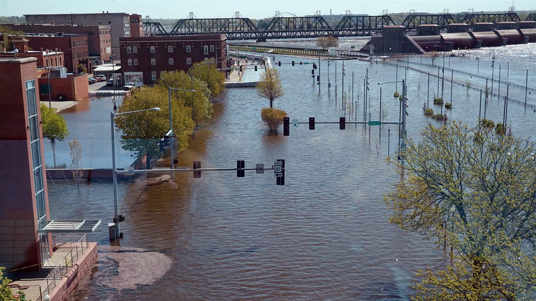 Floodwater covers the streets and other low-laying surfaces in an urban area, which includes several multistory commercial buildings. The center of the photo looks down a flooded street with traffic lights standing above the water; in the distance, a truss bridge crosses a waterway. 