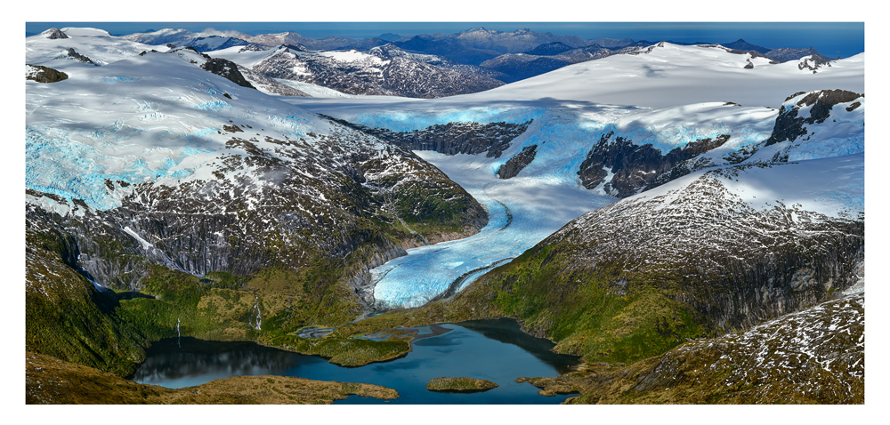 An aerial shot shows a massive, blue-tinted glacier bordered by green mountains that flow down to the glassy, calm waters of a coastal cove. 