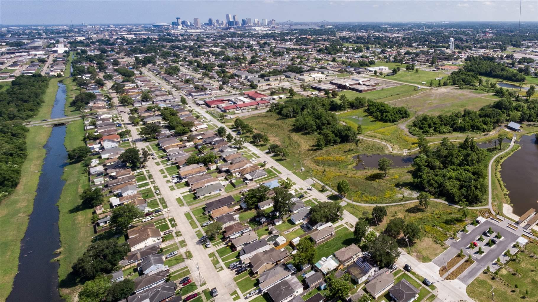 An aerial view shows a residential neighborhood with a waterway on the left and a large park, with a lake, trees, and several grassy areas, to the right. Developed areas surround the other sides of the park, and a city skyline rises in the distance.