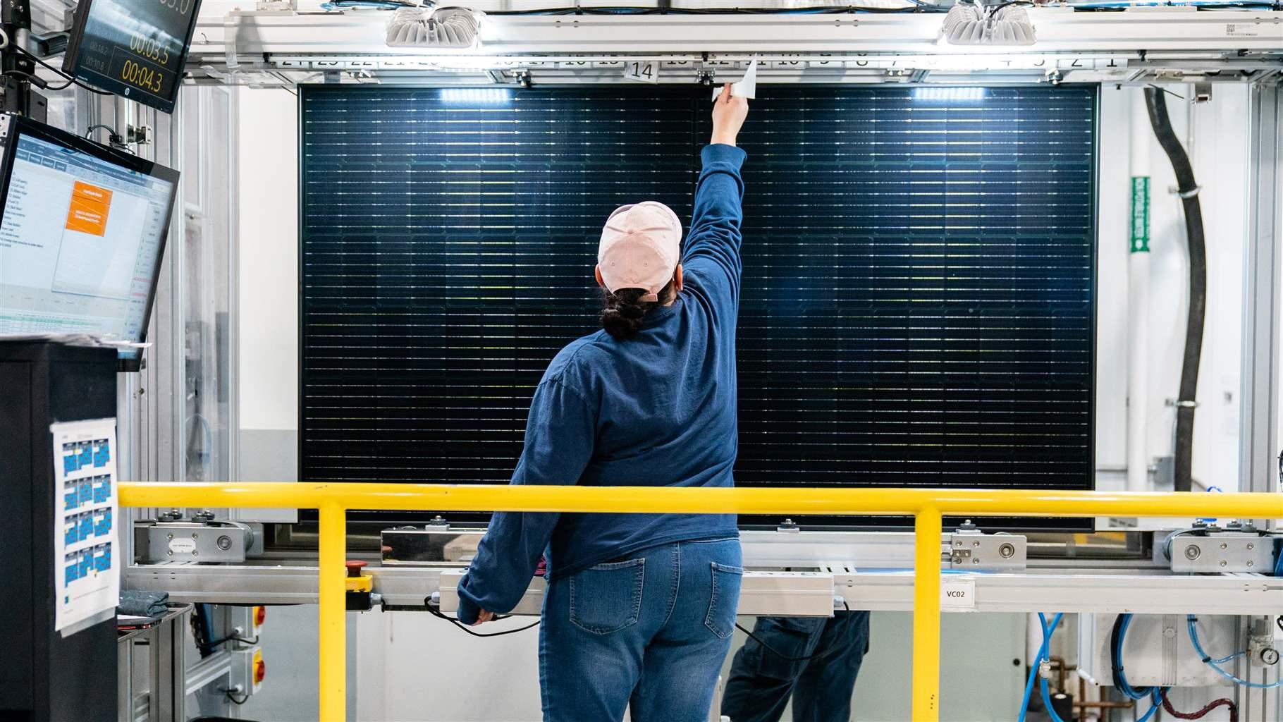 A person in a cap with their back to the camera faces a solar panel in a brightly lit industrial setting and reaches up to check the panel.
