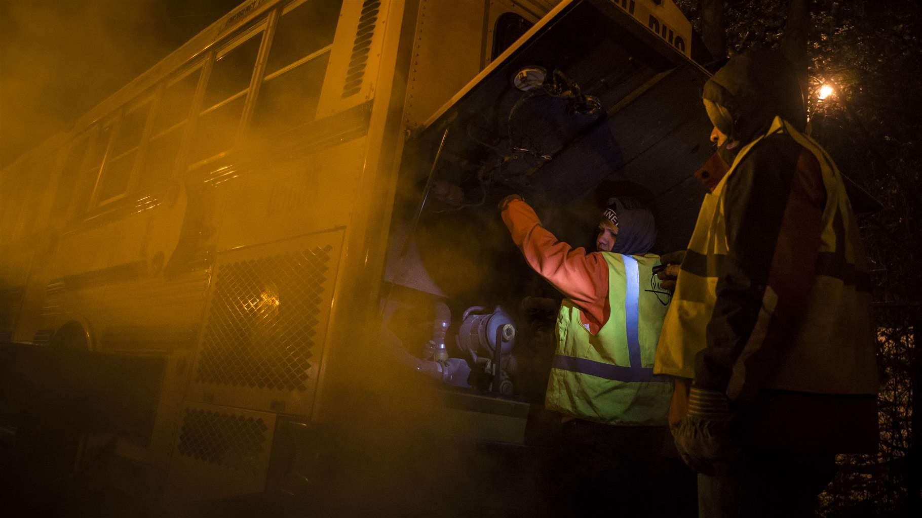 Under amber streetlights, two workers dressed in warm clothing and safety vests work under a flap at the back of a school bus to start it on a dark morning. Bus exhaust is rising from the vehicle adjacent to the workers. 