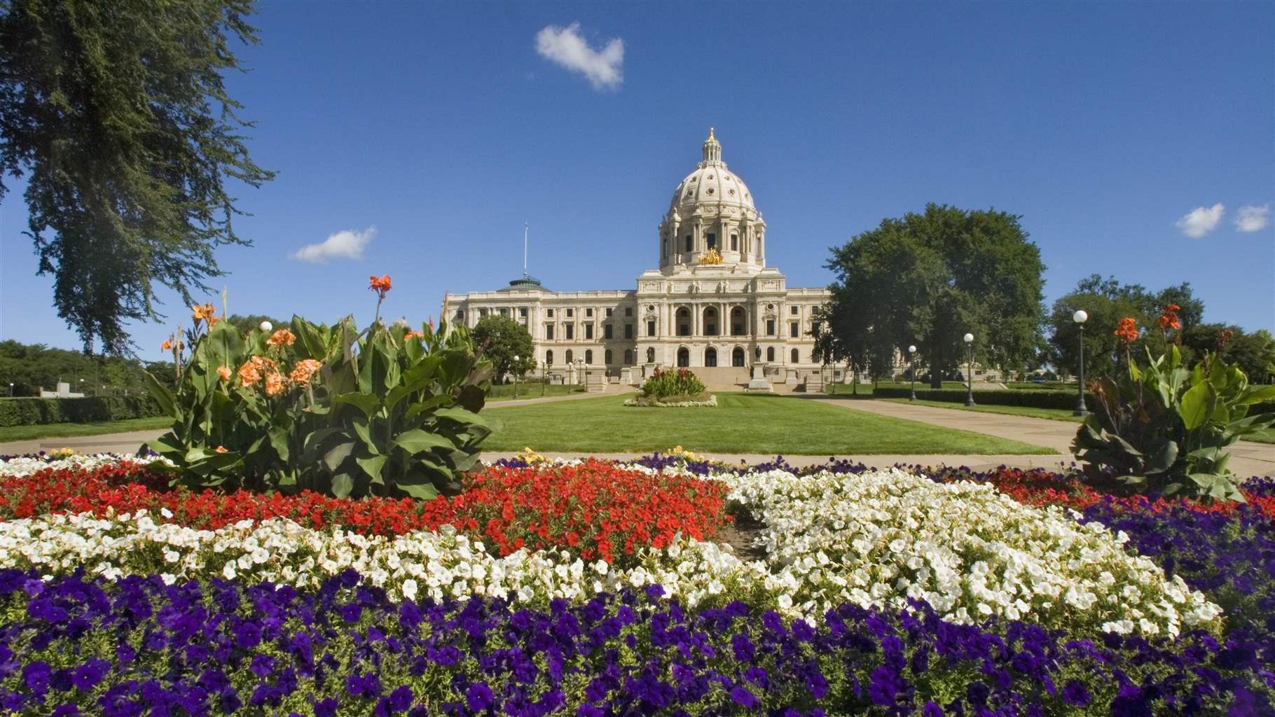A blue sky with a few white puffy clouds sits over a flowerbed filled with red, white, and blue flowers in the foreground. In the background, a green lawn leads to a majestic off-white statehouse building that has a dome in the center.