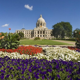 A blue sky with a few white puffy clouds sits over a flowerbed filled with red, white, and blue flowers in the foreground. In the background, a green lawn leads to a majestic off-white statehouse building that has a dome in the center.