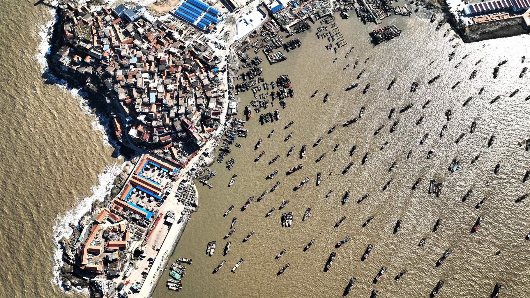 An aerial shot of dozens of fishing boats in brown waters at a harbor in China’s Jiangsu Province. Many buildings are clustered together on a sandy outcropping that houses the port.