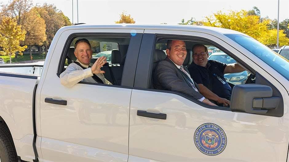 Three smiling people sit in a parked white pickup truck with a blue state seal on the passenger side door. From the back passenger seat, a person waves to the camera through the open window. In the front two seats, the passenger and the driver—who has one hand on the steering wheel—look out the passenger window toward the camera.