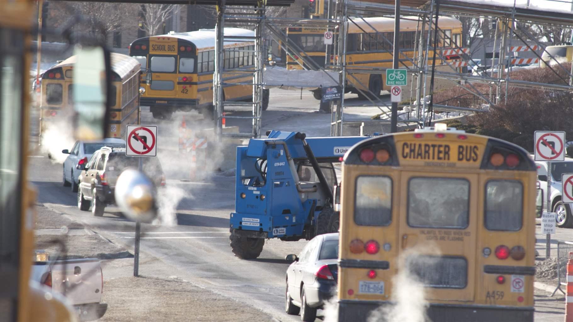 Four yellow school buses emit smoke as they travel a road crowded with cars, construction vehicles, and scaffolding.  
