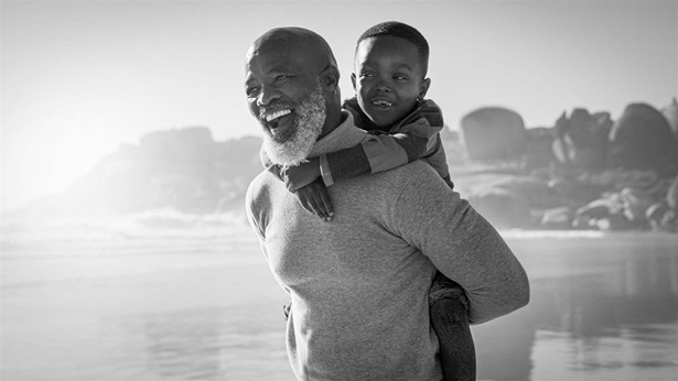 A black and white photo showing a person with a gray beard, carrying a child on his back as they walk along a beach. Both look into the distance ahead, smiling wide.