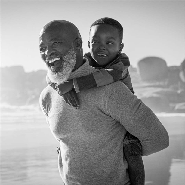 A black and white photo showing a person with a gray beard, carrying a child on his back as they walk along a beach. Both look into the distance ahead, smiling wide.