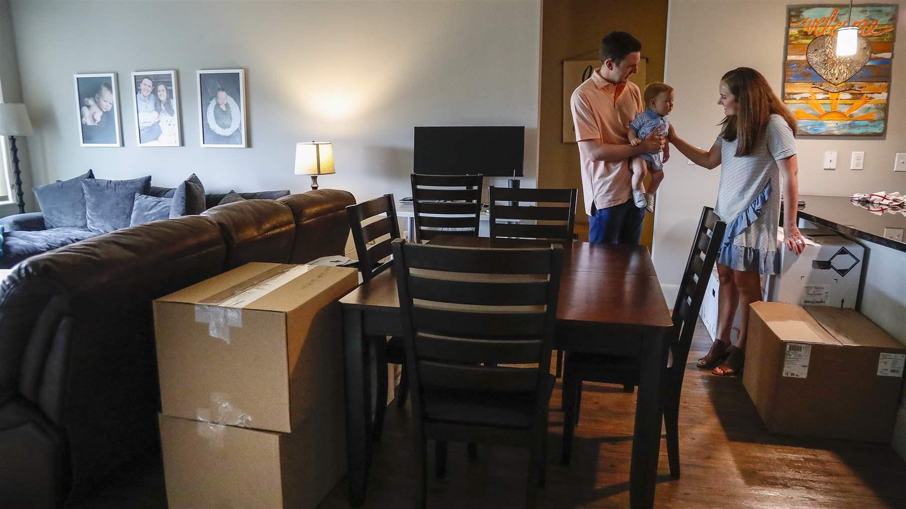 A couple with a baby stand in a living area amid unpacked moving boxes next to a brown dining set. A gray sectional sofa delineates the living room to the left, and a counter indicates a kitchen to the right. Family photos and artwork hang on the walls.