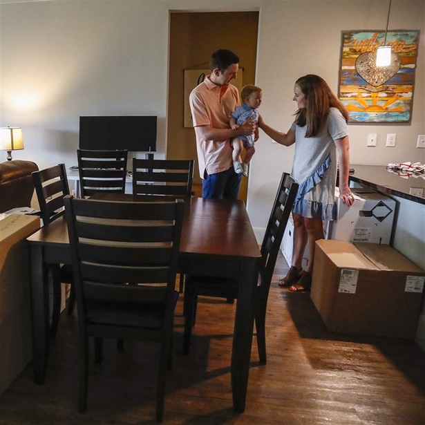 A couple with a baby stand in a living area amid unpacked moving boxes next to a brown dining set. A gray sectional sofa delineates the living room to the left, and a counter indicates a kitchen to the right. Family photos and artwork hang on the walls.