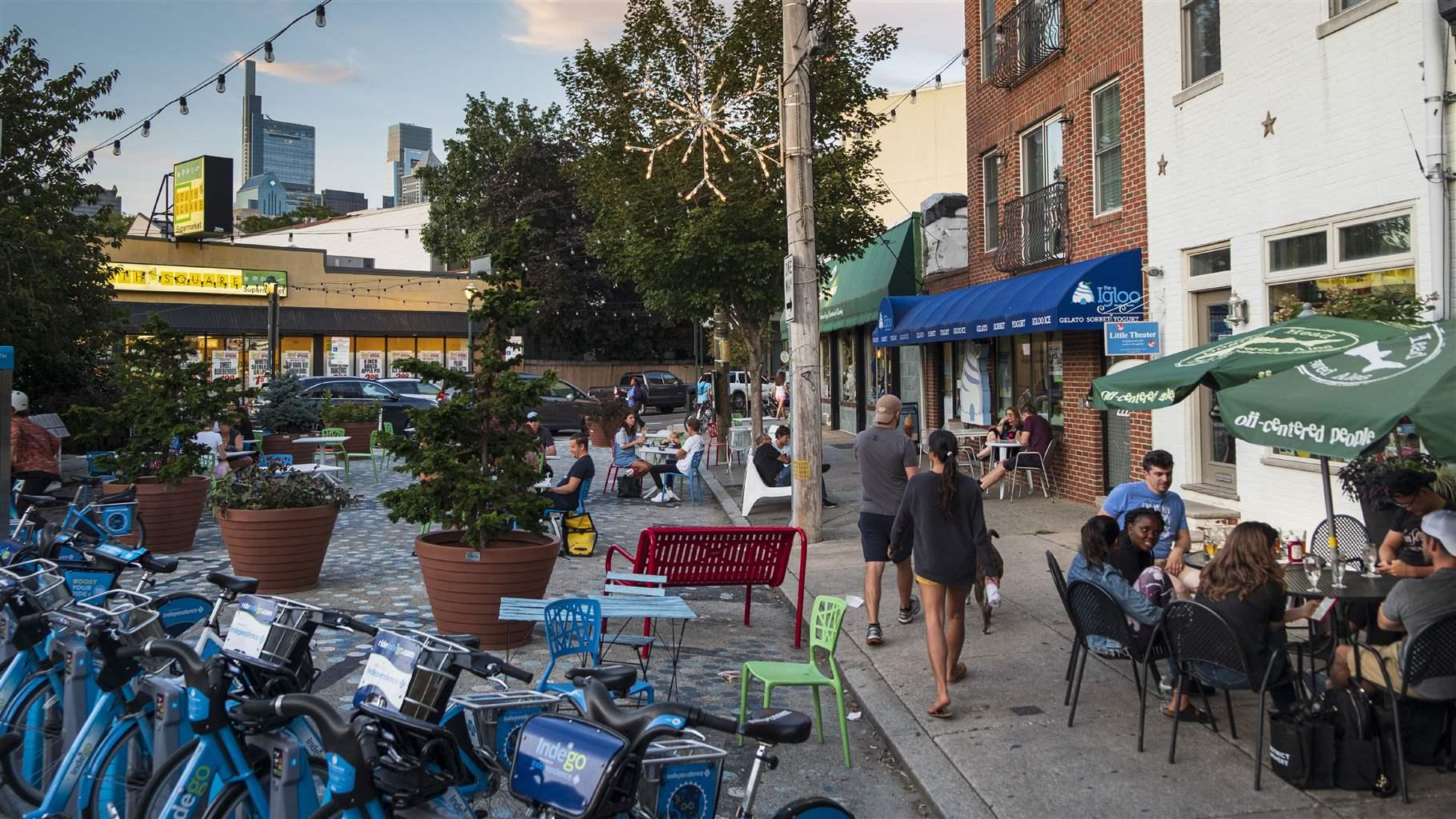 Three groups of diners sit at outdoor tables at restaurants as pedestrians make their way down a busy Philadelphia street. 