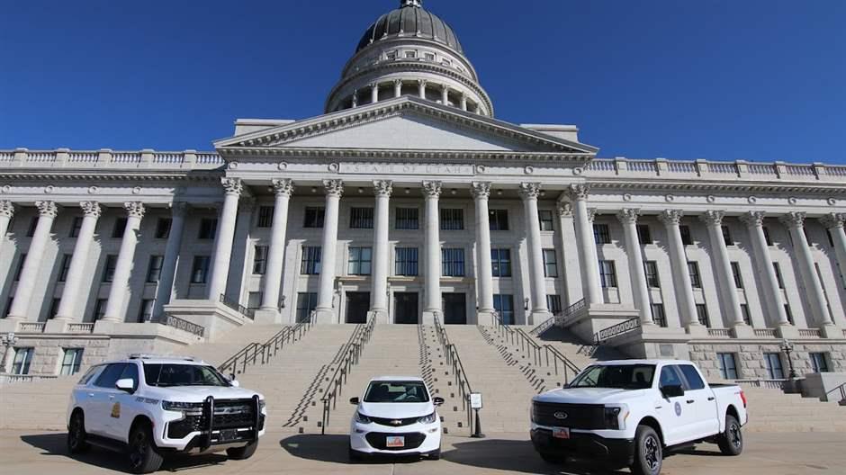 Three white vehicles sit at the base of the Utah state Capitol. Steps flanked with railings rise behind the vehicles and lead to an off-white building adorned with columns across the front.