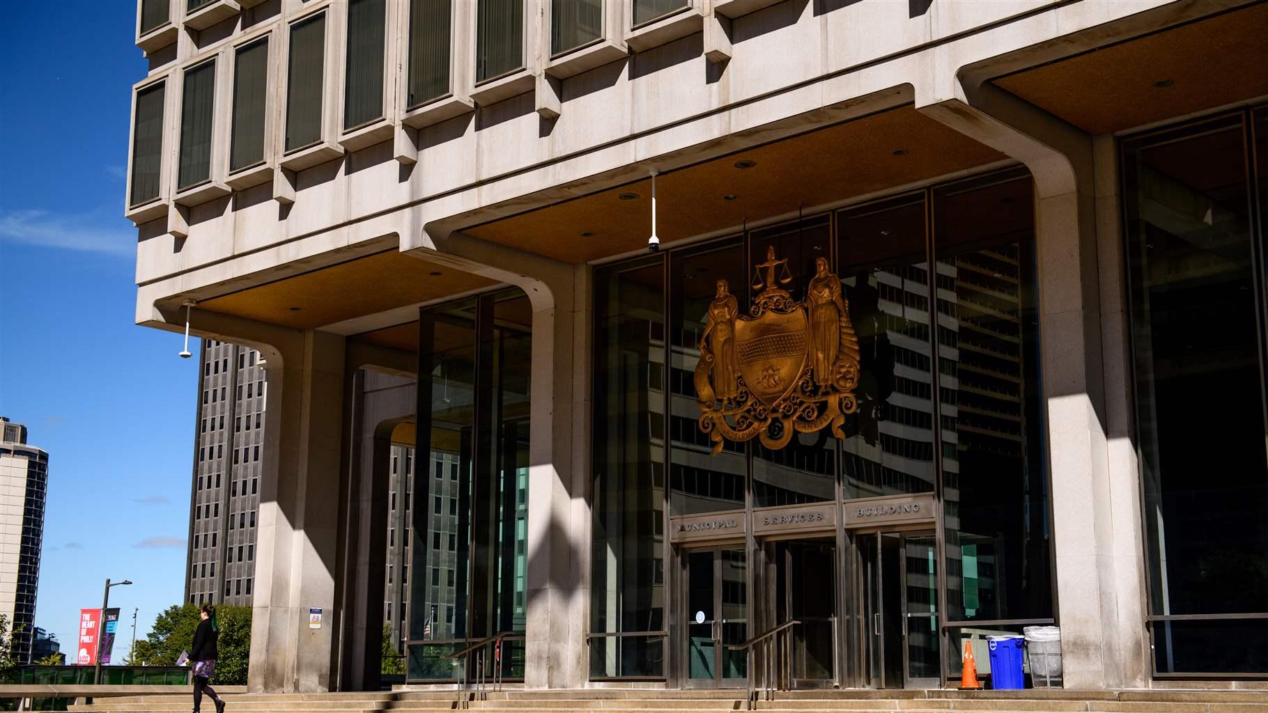 A large, golden city crest appears above the glass doorways to Philadelphia’s Municipal Services Building as a person dressed in a black sweater and purple skirt walks down the concrete steps in front of the building. The crest comprises a shield with two female figures on either side, one holding a wreath and the other a cornucopia. An arm rises from the top of the shield holding a scale of justice. The words “Philadelphia Maneto” appear at the bottom, meaning “Let Brotherly Love Continue.”