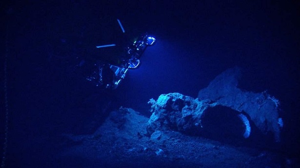 An underwater robot glows blue against a dark background as it shines a light illuminating several large rocks on the sea floor.